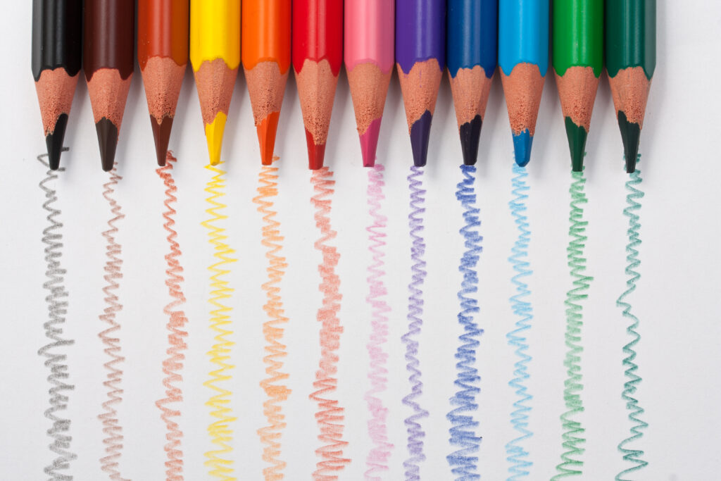 A rainbow row of colored pencils over a sheet of paper, each one of them drawing a wiggly line showcasing their bright colors.