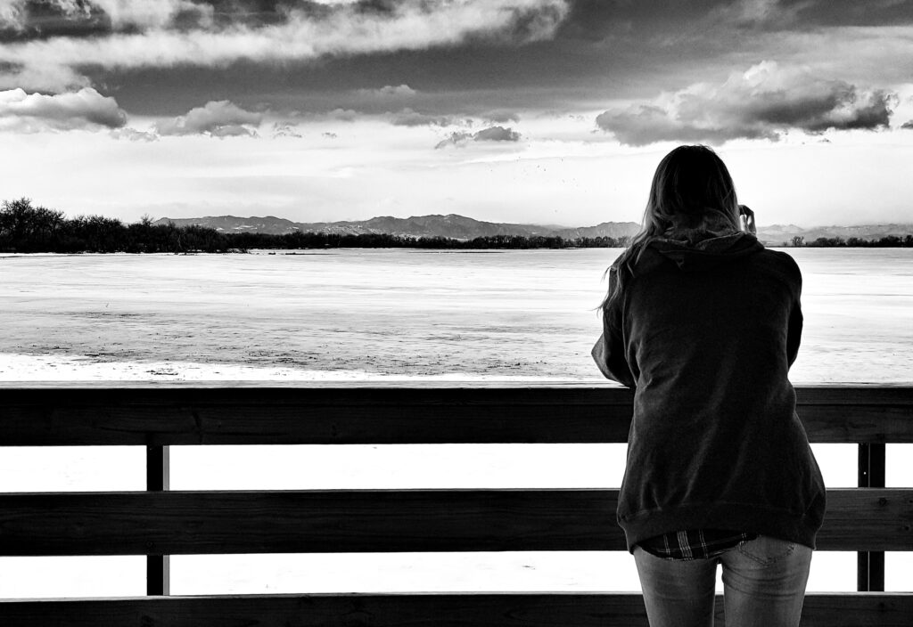 A black and white photo of a person with long hair wearing a hoodie, leaning on a wooden railing and gazing out at a vast, frozen lake. The scene is quiet and contemplative, with dramatic clouds hanging low in the sky and distant mountains barely visible on the horizon. The person stands alone, facing away from the camera, evoking a sense of solitude and reflection.