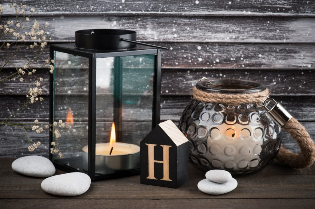 A cozy, rustic tabletop arrangement featuring two lit candles: one inside a black metal lantern with glass panels, and another inside a round, textured glass jar with a thick rope handle. White pebbles are scattered around the candles, along with a small wooden house-shaped block painted black with a large gold "H" on the front. Delicate dried flowers lean gently into the frame from the left. The background is a weathered wooden wall dusted with white speckles, evoking a wintry or snowy feel.